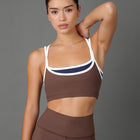 A woman modeling a matching brown activewear set consisting of a sports bra with white and navy trim and high-waisted leggings, set against a solid gray studio background.