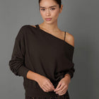 A woman modeling a dark brown off-the-shoulder long-sleeve sweatshirt with a thin black underband and black leopard-print leggings, set against a solid gray studio background.