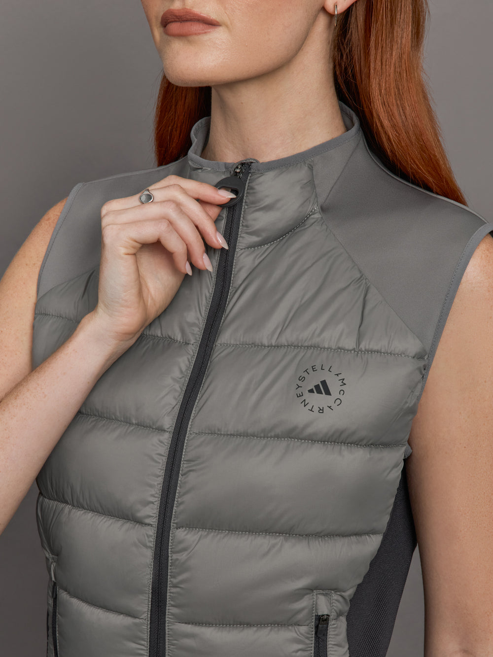 Model wears sleeveless grey golf vest with a high collar, front zipper, and side zip pockets.
