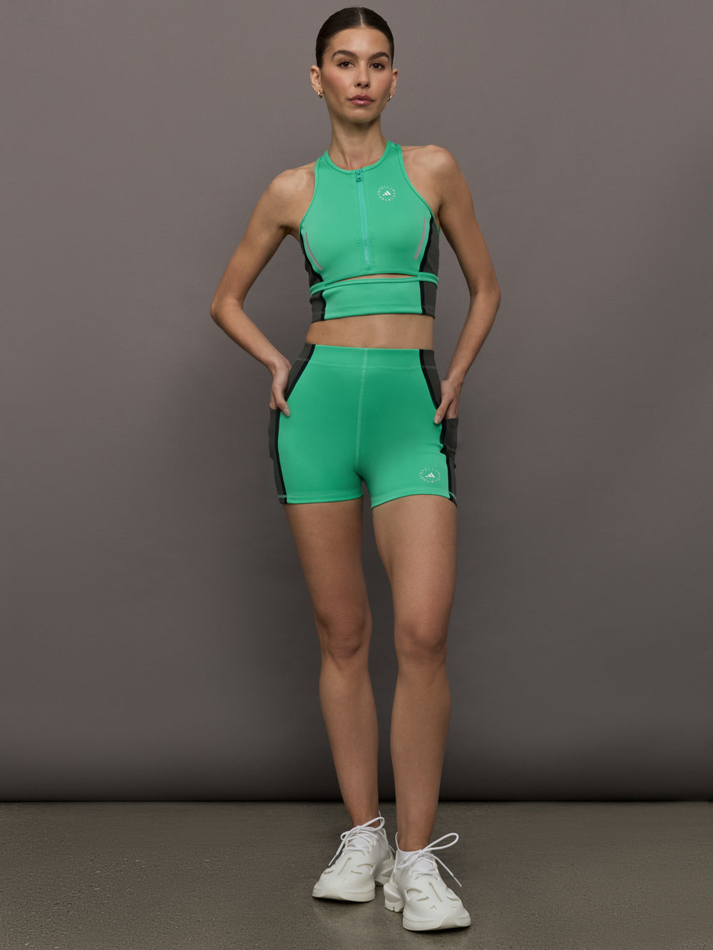 Model wears a bright green cropped sleeveless activewear tank with gray side paneling and an open horizontal slit below the chest. 