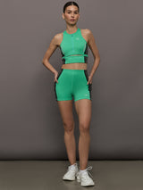 Model wears a bright green cropped sleeveless activewear tank with gray side paneling and an open horizontal slit below the chest. 