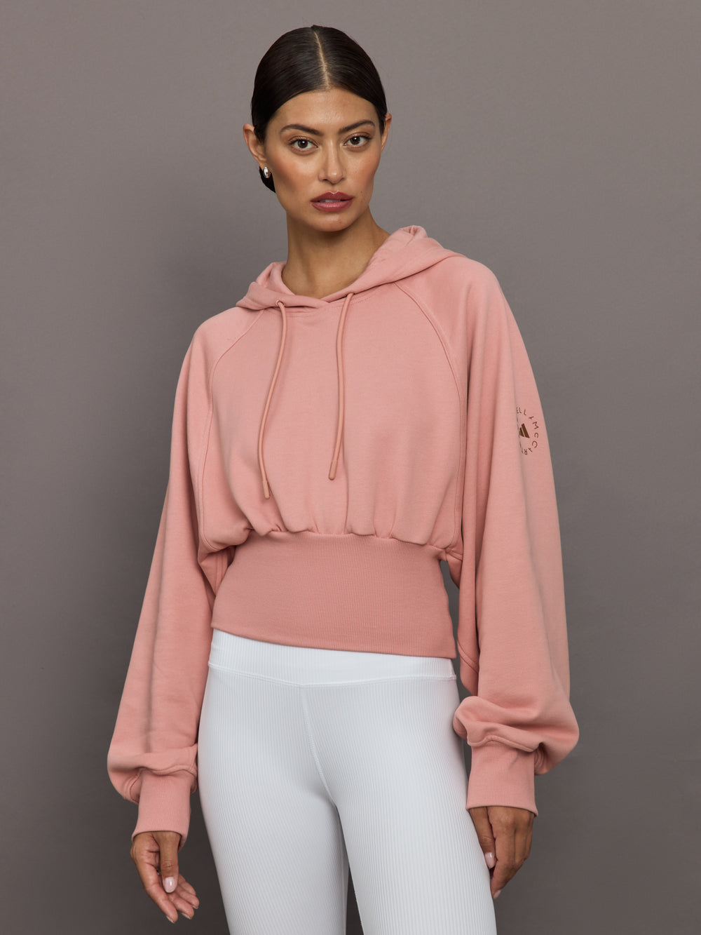 Woman wearing a pink hoodie and white pants against a gray background
