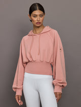 Woman wearing a pink hoodie and white pants against a gray background