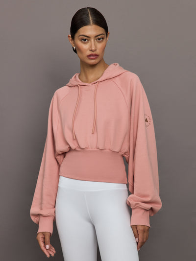 Woman wearing a pink hoodie and white pants against a gray background