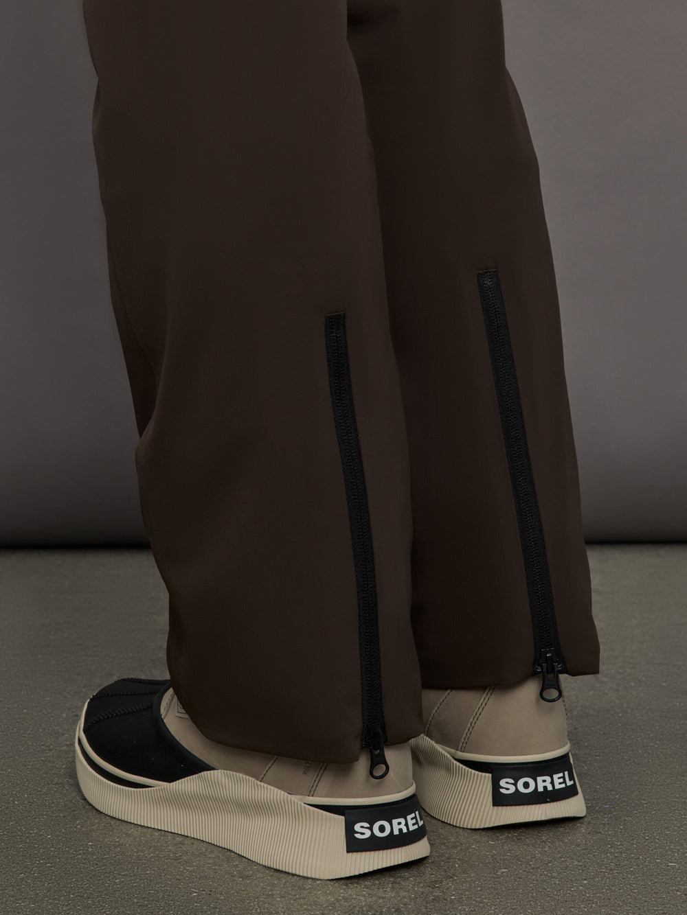 Brown pants with zippers and beige sneakers with SOREL logo on a gray background