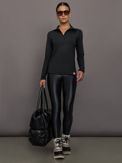 Person wearing a black long-sleeve top and black leggings, holding a black bag against a gray background.