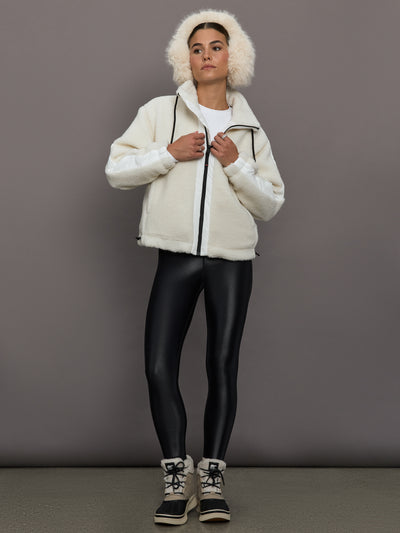 Person wearing a white jacket with fur hood, black leggings, and black boots on a gray background