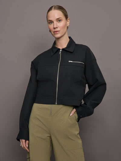 Model wears a collared cropped black bomber jacket with silver hardware and a horizontal pocket over left chest with zipper. 