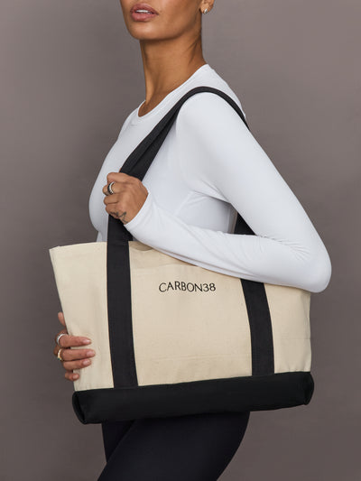 Model is carrying Carbon38 branded tote bag in ivory canvas with black straps and base, side view.