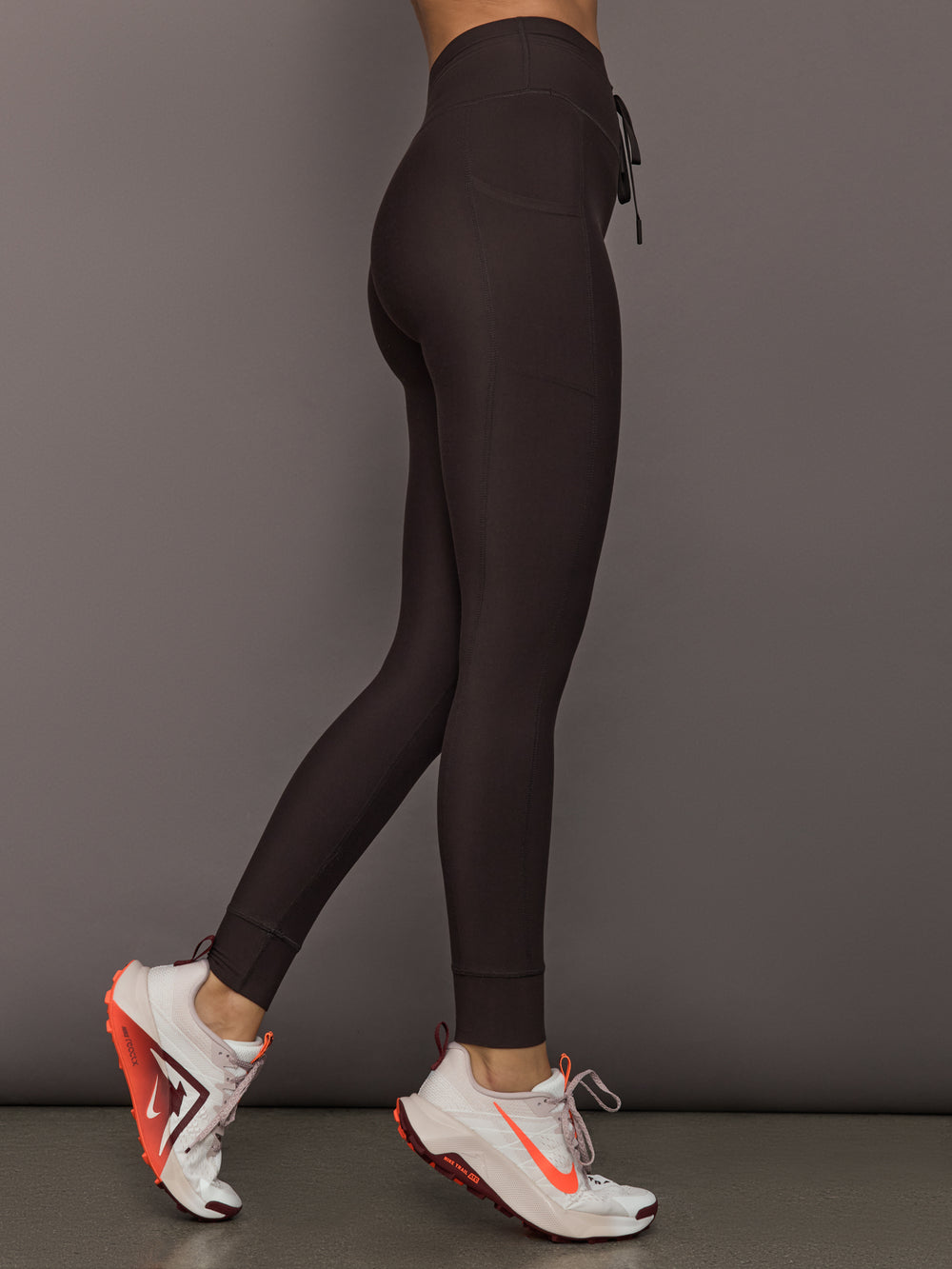 Model is wearing dark brown high-rise leggings with a drawstring waistband and slim side pocket, full-length compression fit, side view.
