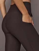 Model is wearing dark brown high-rise sport leggings with a wide waistband and contoured seam detailing, full-length fit, close-up back view.