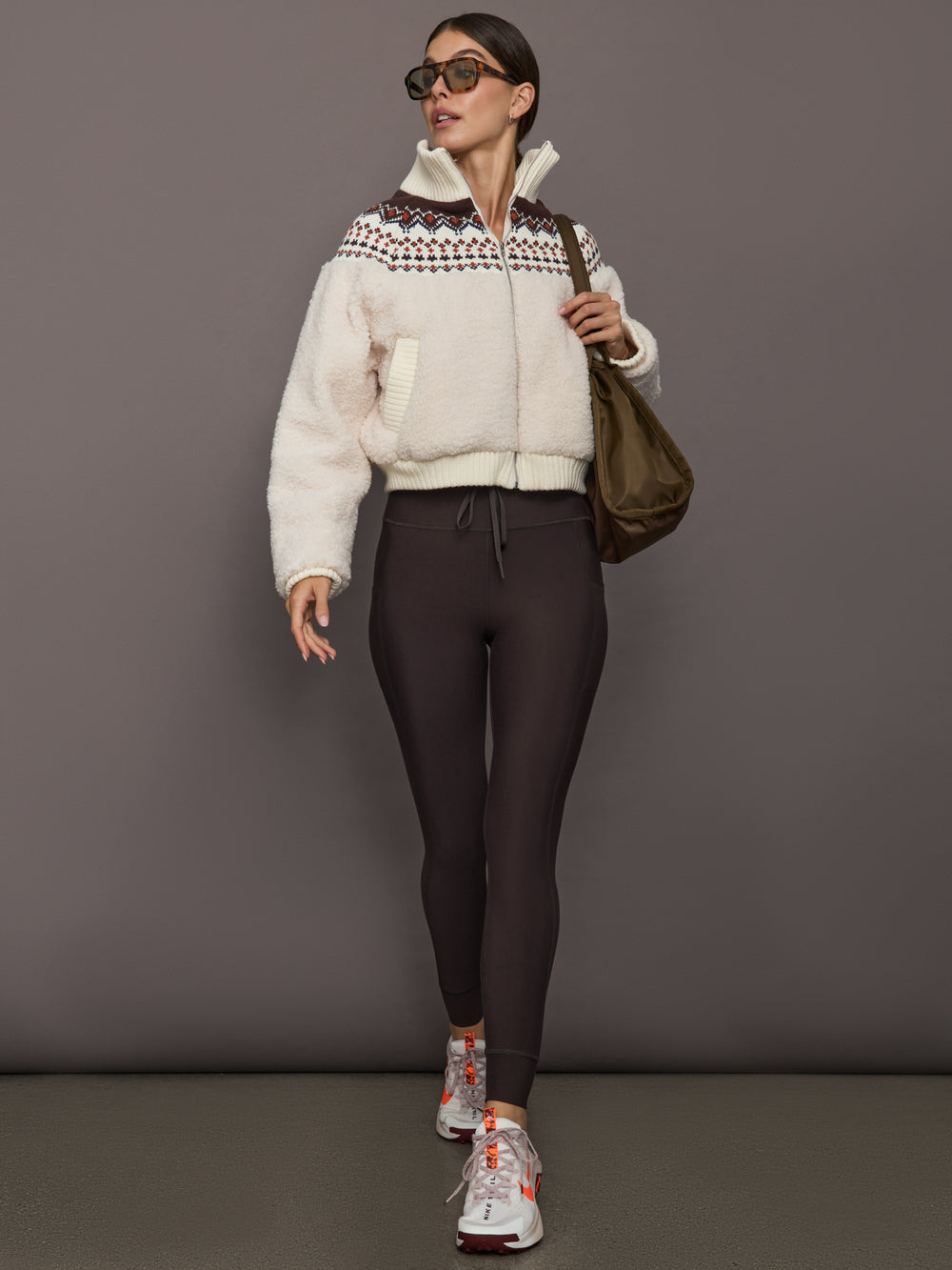 Model in dark brown high-rise leggings with drawstring waist and pockets, paired with a cream sherpa zip-up jacket, oversized sunglasses, brown shoulder bag, and sneakers, front view.