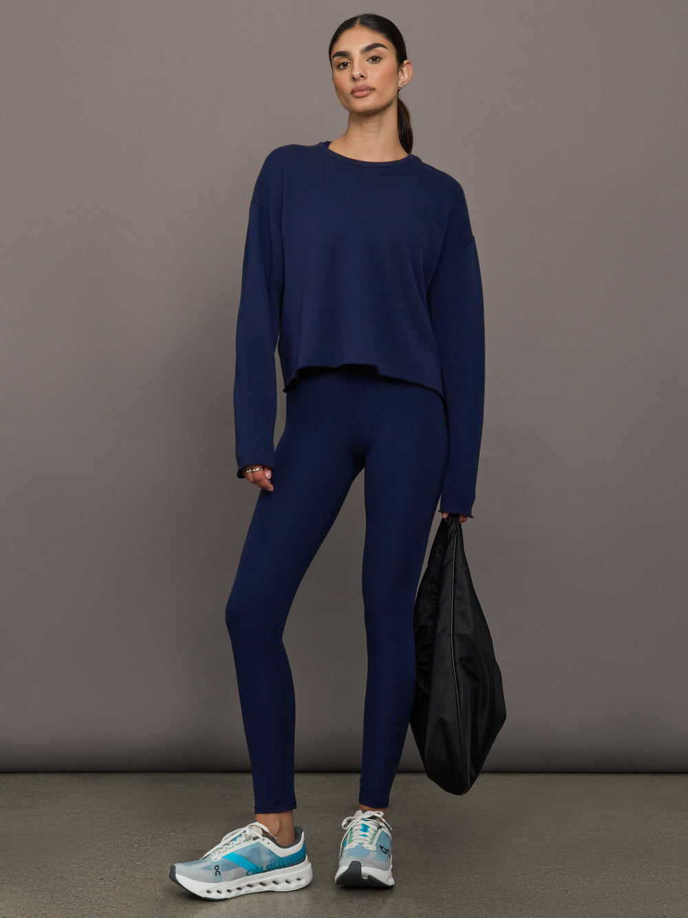 Woman wearing a navy blue outfit holding a black bag against a gray background