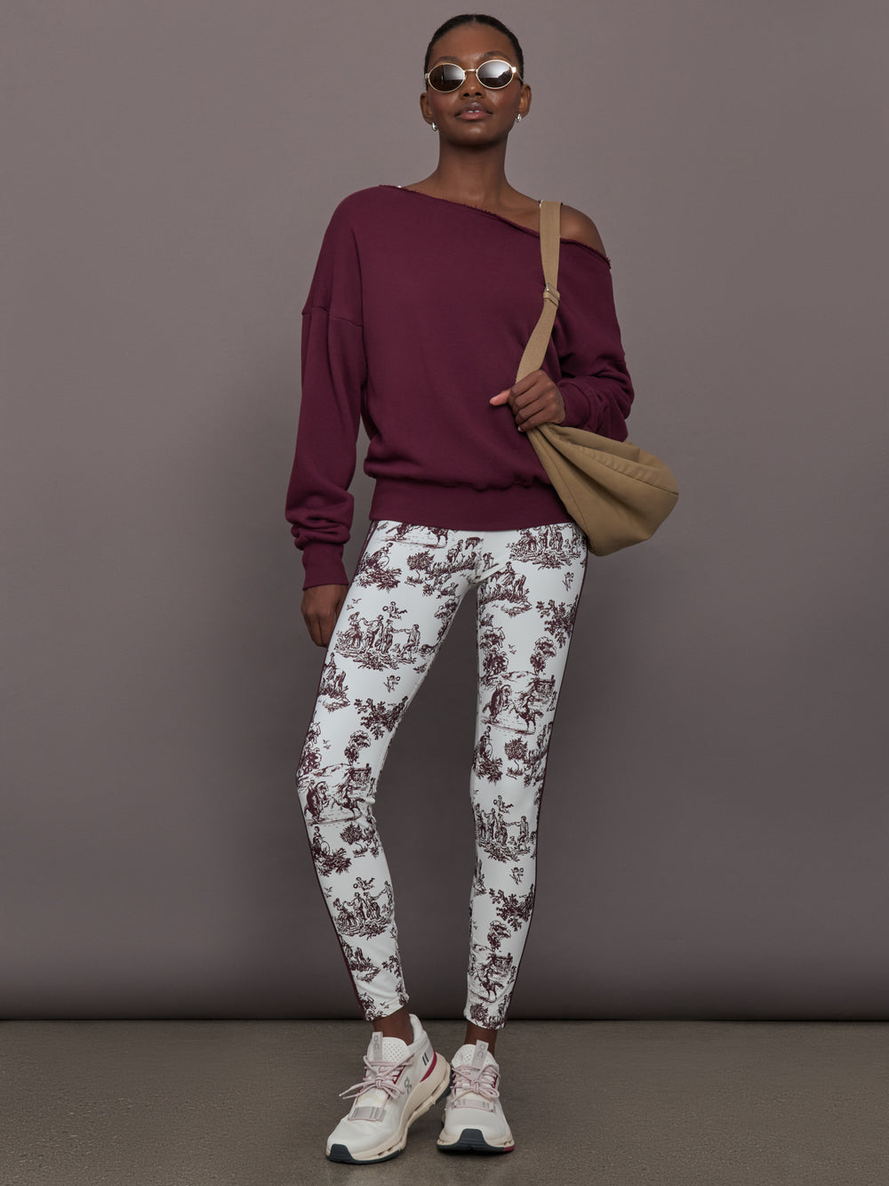 Woman wearing a burgundy off-shoulder top and white toile print pants, holding a beige bag against a gray background.