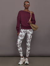 Woman wearing a burgundy off-shoulder top and white toile print pants, holding a beige bag against a gray background.