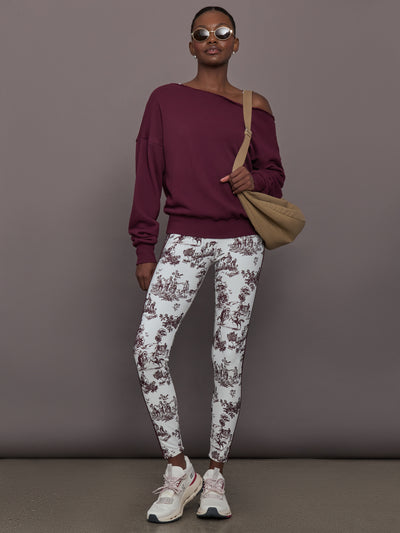 Woman wearing a burgundy off-shoulder top and white toile print pants, holding a beige bag against a gray background.