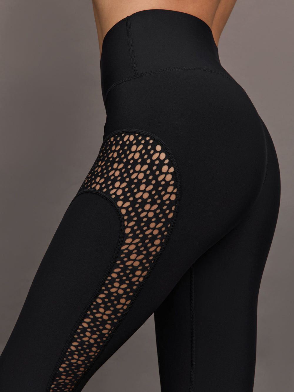 Model wears a pair of black high waisted leggings with laser cutouts along thighs. 