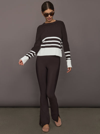 Woman wearing a brown and white striped sweater and brown leggings against a gray background, full body shot. 