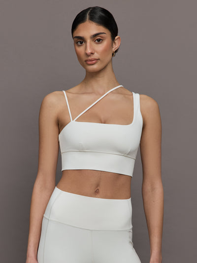 Model wears a white sports bra with one thick strap and two asymmetrical spaghetti straps. 