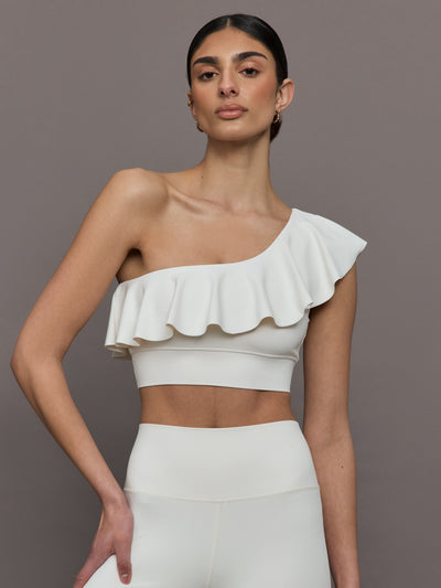 Model wears a white one shoulder sports bra with a ruffled overlay. 