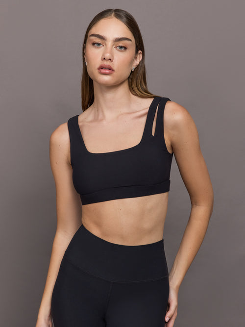 Model wears Black sports bra with double straps on one side and a square neck.