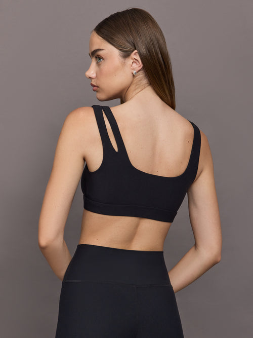 Model wears Black sports bra with double straps on one side and a square neck.