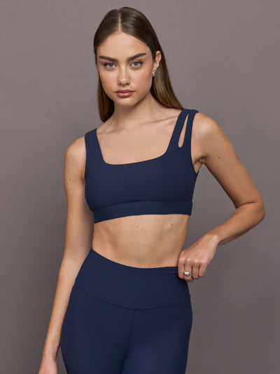 Model wears Navy sports bra with double straps on one side and a square neck.