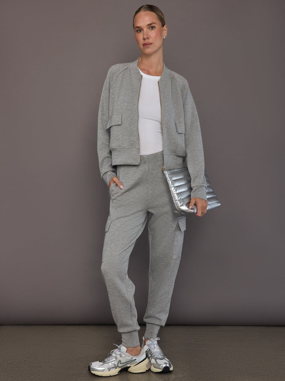 Model is wearing a matching set in heather grey ultrasoft fabric featuring the bomber jacket and cargo jogger, styled with a white ribbed tank top, silver sneakers, and a metallic clutch, full front view.