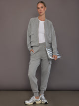 Model is wearing a matching set in heather grey ultrasoft fabric featuring the bomber jacket and cargo jogger, styled with a white ribbed tank top, silver sneakers, and a metallic clutch, full front view.