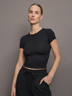 Model wears a short sleeved, ribbed jersey fitted black top with ruching down both sides. 