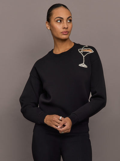 Model wearing black martini embellished sweatshirt with crystal martini glass detail on shoulder, front close-up view.