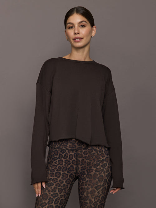 Model is wearing a crew sweatshirt in dark brown with dropped shoulders and raw straight hemline, front view.