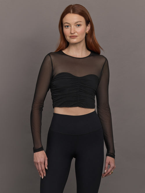 Model wears the The Melt and Mesh Ruched Top in Black, with a ruched center front, mesh on the sleeves and upper chest, and is cropped.