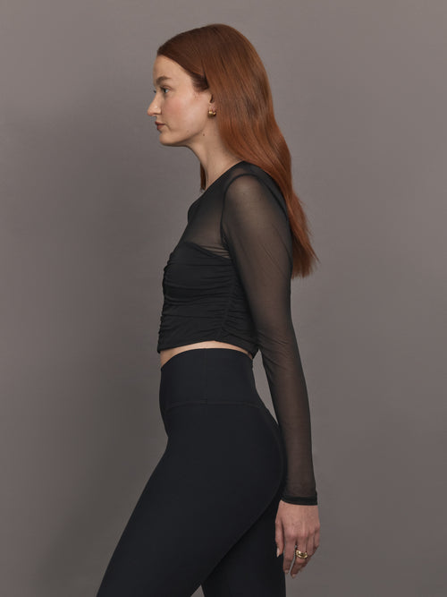 Model wears the The Melt and Mesh Ruched Top in Black, with a ruched center front, mesh on the sleeves and upper chest, and is cropped.