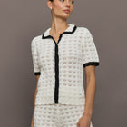 Model wears white and black crochet button-down top with short sleeves and a collared neck.