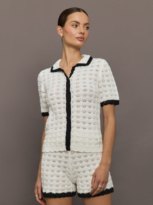 Model wears white and black crochet button-down top with short sleeves and a collared neck.