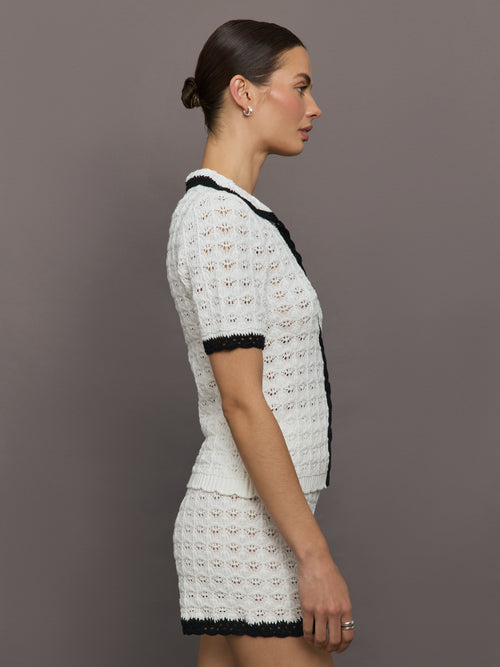 Model wears white and black crochet button-down top with short sleeves and a collared neck.