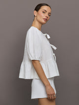 Model wears white puff sleeve top with a front ties and high neckline.
