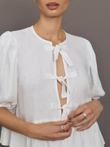 Model wears white puff sleeve top with a front ties and high neckline.

