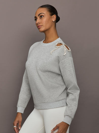Model wearing grey bow embellished sweatshirt with crystal bow detail on shoulder, styled with white leggings, angled side view.