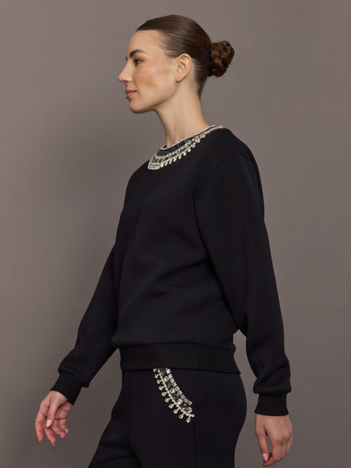 Model in black jewel embellished sweatshirt showing crystal neckline and pocket detail, side view.