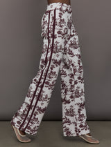 Model wears white track pants with burgundy toile print. 