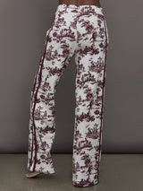 Model wears white track pants with burgundy toile print. 