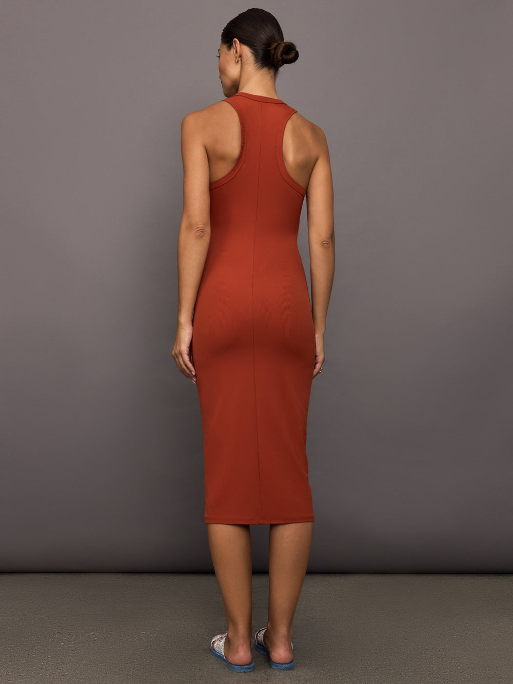 Model wears terracotta-colored, ribbed tank dress with a high neckline, sleeveless design, and a slim, body-hugging fit that extends right below the knee.