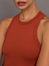 Model wears terracotta-colored, ribbed tank dress with a high neckline, sleeveless design, and a slim, body-hugging fit that extends right below the knee.