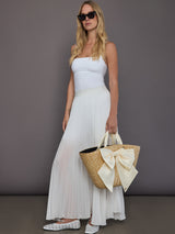 Full-body model view wearing white tank and pleated maxi skirt styled with oversized straw tote featuring ivory bow.