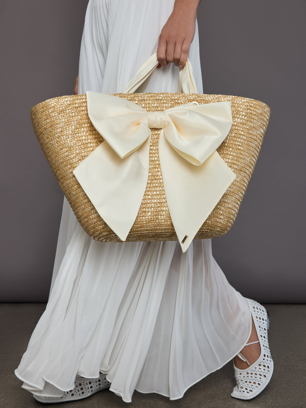 Model holding oversized straw tote with ivory bow styled with white pleated maxi dress and cutout flats.