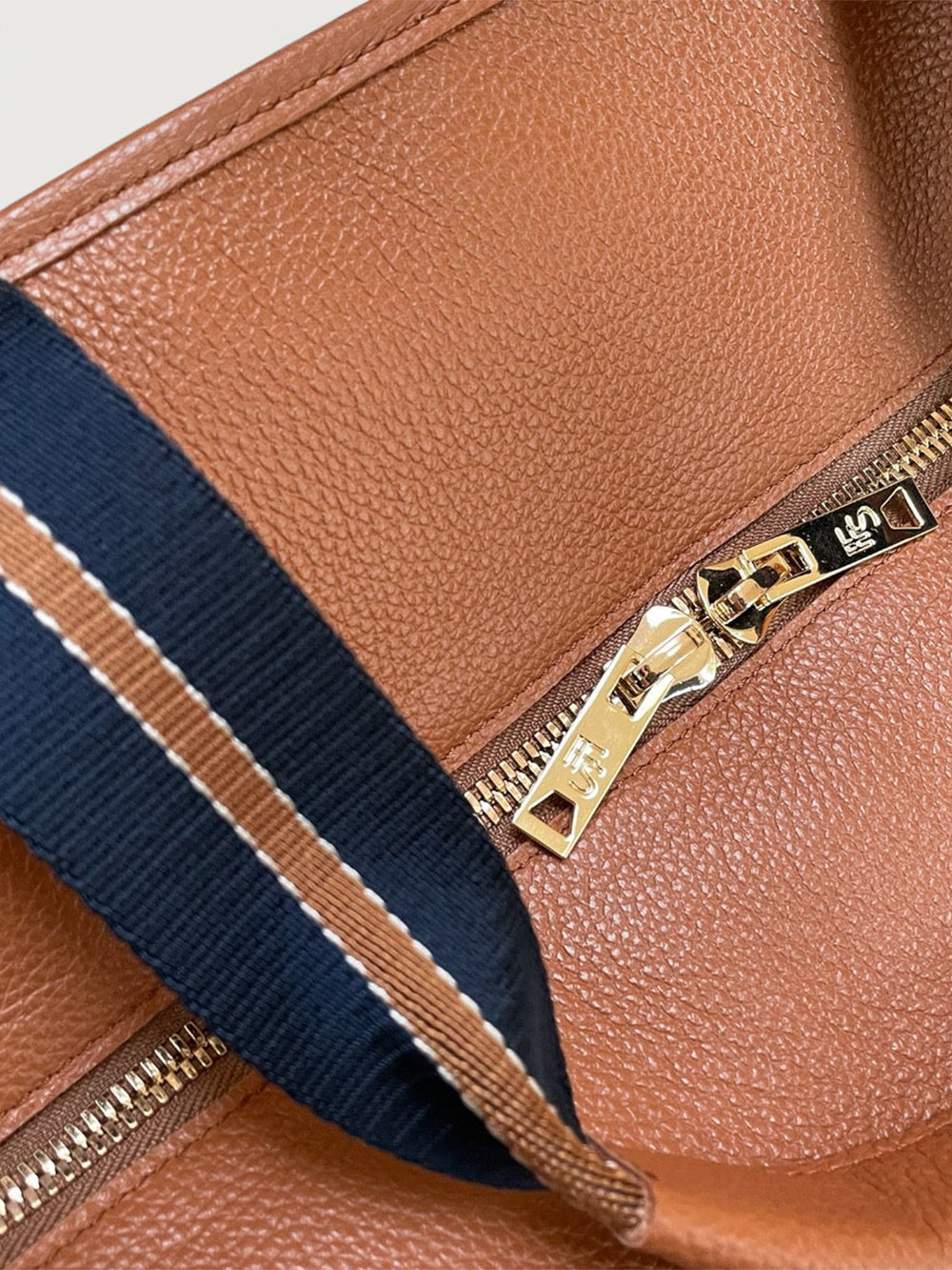 Close-up of tan leather tote bag with gold double zipper pull and navy striped strap.