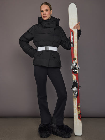 Model wearing the Bufera Ski Jacket in black, full body view holding skis with the white belt visible.