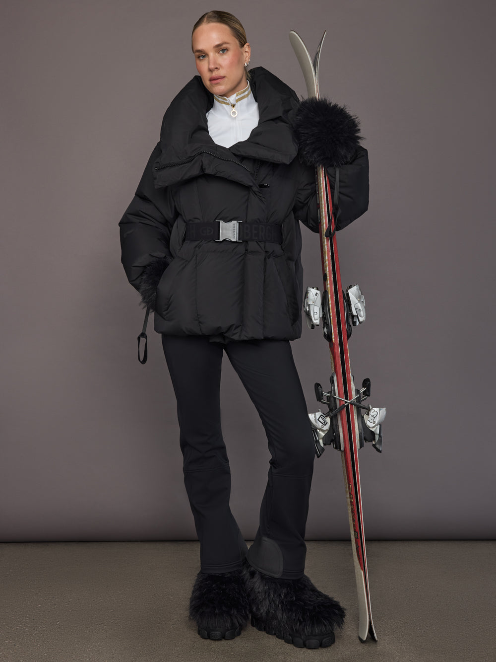 Black quilted down jacket on model, full body ski outfit with belt.